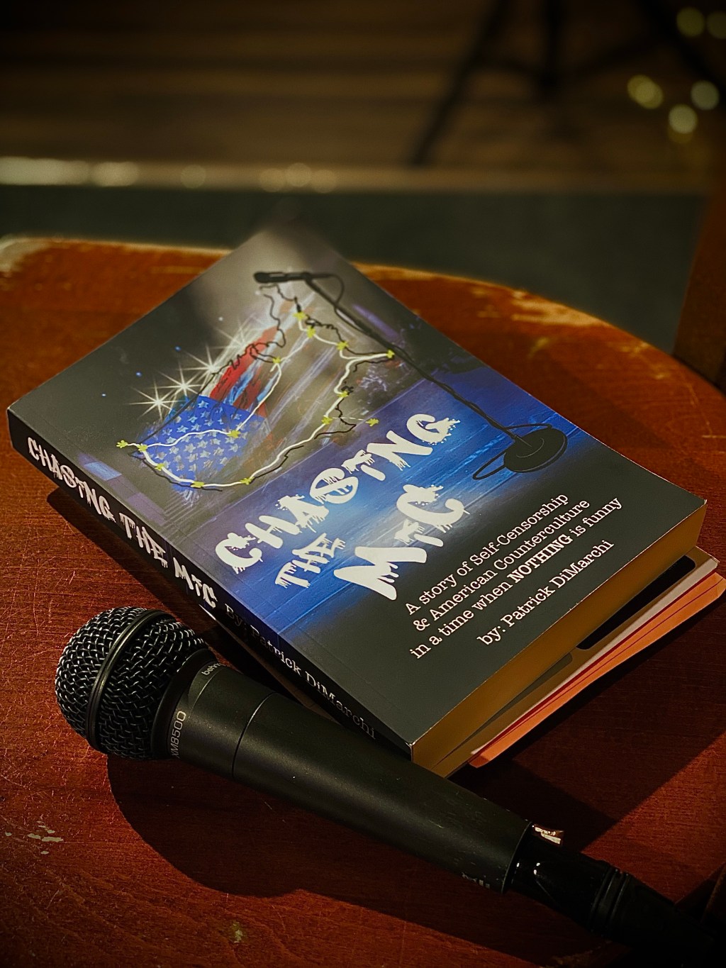 A picture of a book sitting on stool next to a microphone at a stand-up comedy open mic night. The title of the book is Chasing The Mic.